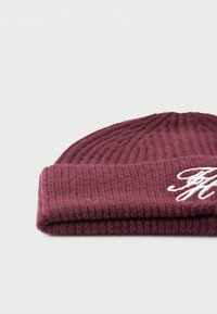 Foldet vinrød strikket hue med hvide broderede initialer på kanten, vist på en ensfarvet hvid baggrund.