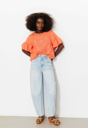 Femme aux cheveux bouclés et volumineux portant un chemisier orange à manches volantées, un jean taille haute bleu clair et des sandales compensées beige.