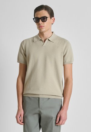 Jeune homme portant un polo beige texturé à manches courtes, pantalon gris et lunettes de soleil noires, debout devant un fond uni.