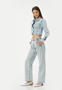 Chaqueta de mezclilla recortada en azul claro y jeans de tiro alto con un corte holgado, que cuentan con detalles de tachuelas y bolsillos laterales. Zapatos planos blancos.