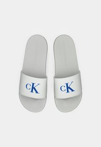 Fehér Calvin Klein fürdőpapucs kék "cK" logóval a pántokon, felülről nézve, sima fehér háttérrel.