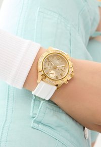 Montre dorée avec un cadran texturé, deux sous-cadrans et des accents en cristal ; bracelet en caoutchouc blanc ; portée sur un vêtement bleu clair.