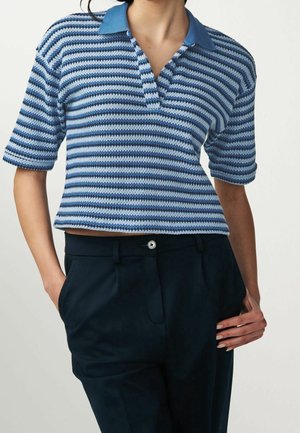 Personne portant un haut tricoté à manches courtes rayé bleu avec col et un pantalon bleu foncé, une main dans la poche et l'autre reposant sur la hanche.