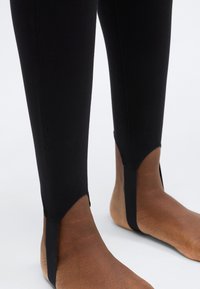 Picioare de costum de scufundare negru cu finisaj neted, prevăzute cu secțiuni decupate la nivelul gleznelor, care lasă pielea la vedere și asigură flexibilitate. Detaliu subtil de cusătură vizibil.