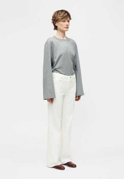 Personne debout, portant une chemise à manches longues grise, un pantalon large blanc taille haute et des chaussures marron, devant un fond blanc uni.