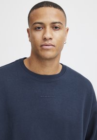 Marineblauwe crewneck sweatshirt van zachte stof, met een kleine tekstgraphic "WAARIN BEN JIJ RIJK?" op de borst en ribbelboord bij de hals.