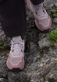 Primo piano di piedi che indossano scarpe da ginnastica viola chiaro, in piedi su una superficie di pietra grigia ruvida con chiazze di muschio verde.