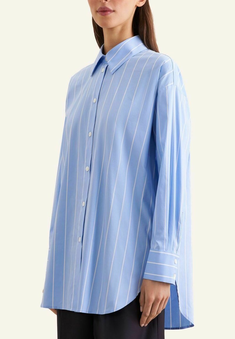 Femme portant une chemise bleue ample à manches longues avec des rayures verticales blanches et des boutons blancs, associée à un pantalon noir.