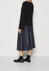 Haut à manches longues noir associé à une jupe midi en cuir plissé bleu foncé. Chaussures noires texturées mises en valeur par des chaussettes noires.