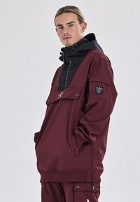 Bordeaux farvet pulloverjakke med en sort hætte, der har en brystlomme, snor i hætten og et logo-patch på ærmet, lavet af struktureret stof.