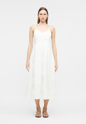 Frau trägt ein weißes Midikleid mit dünnen Trägern, Blumenstickerei und hellbraunen Riemchensandalen, steht vor einem schlichten hellen Hintergrund.