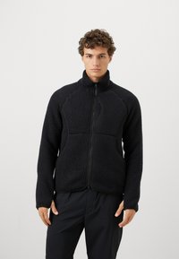 Giovane uomo che indossa una giacca in pile nera con zip e colletto alto e pantaloni neri, in piedi contro uno sfondo grigio chiaro uniforme.