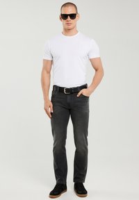 Férfi fekete napszemüvegben, fehér pólóban, fekete övvel, sötétszürke farmerben és fekete tornacipőben, egyik kezével a zsebében áll.