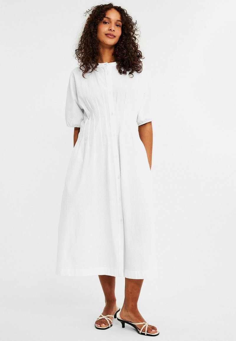 White midi dress with short puff sleeves, textured fabric, button front, and gathered waist. Worn with black and beige strappy sandals.