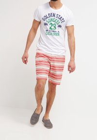 Bílá tričko s barevným textem a grafikou, červené a bílé proužkované šortky, šedé slip-on boty. Model stojí proti neutrálnímu pozadí.