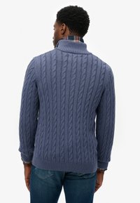 Sweter w splot warkoczowy w stonowanym niebieskim kolorze z wysokim kołnierzem, ściągaczami na rękawach i dole. Warstwowo noszony na koszuli w kratę widocznej przy kołnierzu.