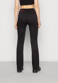 Calças pretas evasé com uma textura suave, apresentando bordados em vermelho nas costas e detalhes de botão nas laterais.