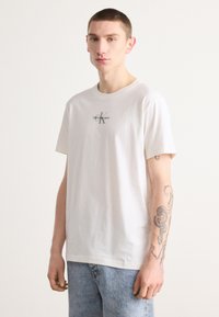 Balta medvilninė marškinė su trumpomis rankovėmis, su mažu pilku "Calvin Klein Jeans" logotipu ant krūtinės. Dėvimi su šviesiai mėlynomis džinsinėmis kelnėmis.