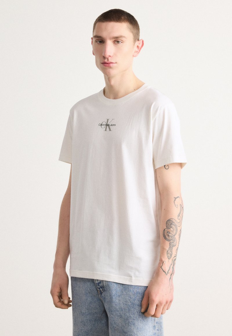 Balta medvilninė marškinė su trumpomis rankovėmis, su mažu pilku "Calvin Klein Jeans" logotipu ant krūtinės. Dėvimi su šviesiai mėlynomis džinsinėmis kelnėmis.