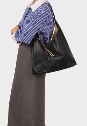 Donna che indossa una camicia a righe blu e una gonna grigia, che porta una grande borsa a tracolla nera in pelle con nappe decorative.