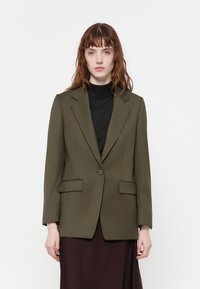 Olivově zelený blazer s klopami, na jedno knoflíkové zapínání a dvěma předními kapsami, nošený přes černý rolák. Hladká textura látky.