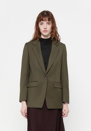 Blazer verde oliva con rever a punta, chiusura a un bottone e due tasche frontali, indossato sopra un dolcevita nero. Tessuto dalla superficie liscia.