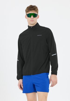 Mand iført sort sportstrøje med lynlås med "Endurance" logo, lyseblå shorts og store grønne reflekterende sportssolbriller.