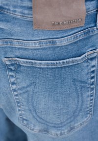 Jeans en denim bleu clair avec une étiquette en cuir estampillée "True Religion". Présente une poche arrière avec un design brodé distinctif.