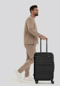 Schwarzer Hartschalenkoffer mit horizontalen Rillen, ausziehbarem Griff und vier Rädern. Eine Person in beigem Outfit zieht den Koffer.