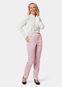 Witte lange mouwen top; lichtroze tapered broek met zijzakken; vrouw in zwarte hoge hakken, staand met hand op de heup.