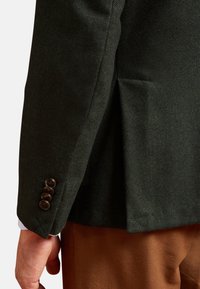Blazer in tessuto a spina di pesce verde scuro con trama testurizzata, dotato di un singolo spacco e quattro bottoni marroni sul polsino. I pantaloni marroni sono visibili.