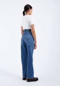 Jeans de denim azul de cintura alta con corte de pierna ancha, que presentan una etiqueta negra en la parte posterior y diseño de cinco bolsillos. Footwear es oscuro y grueso.
