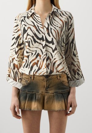 Femme portant une chemise à manches longues à motifs zèbre, boutonnée, rentrée dans une mini-jupe plissée délavée marron et noire.