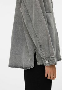 Camicia in denim grigio con vestibilità rilassata, cuciture visibili, polsini con bottone e fessure laterali. Texture liscia e dettagli di design moderni.