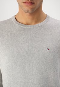 Homme portant un pull gris clair à col rond avec un petit logo rectangulaire rouge, blanc et bleu sur le côté gauche de la poitrine, lèvres et menton visibles.