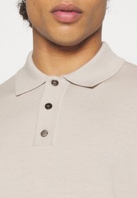Beige polo-tröja med texturerad tyg, med en liten krage och tre mörka knappar. Nära bild som visar halsringningen och knapparna.