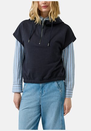 Kobieta w granatowej bluzie z krótkim rękawem z kapturem, założonej na niebiesko-białą koszulę w paski oraz jasnoniebieskie dżinsy.