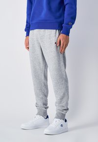 Grå sweatpants med elastiska mudd, sidofickor och en liten logotyp på låret, tillsammans med vita sneakers med blå detaljer.