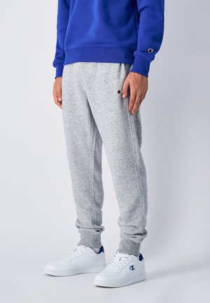 Graue Sweatpants mit elastischen Bündchen, seitlichen Taschen und einem kleinen Logo am Oberschenkel, kombiniert mit weißen Sneakers mit blauen Akzenten.