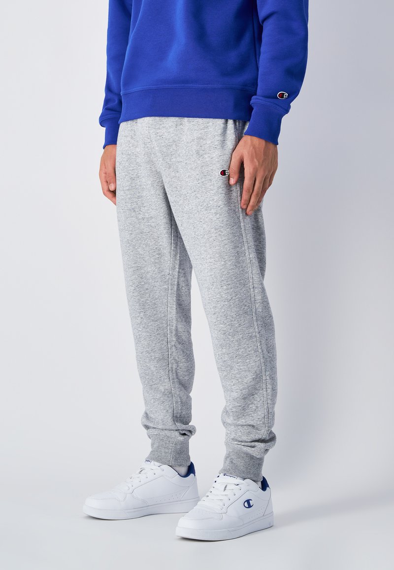 Grå sweatpants med elastiska mudd, sidofickor och en liten logotyp på låret, tillsammans med vita sneakers med blå detaljer.