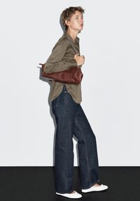 Chemise kaki à manches longues, jean large bleu foncé, chaussures blanches à enfiler et sac à dos en cuir marron. Fond uni, couleurs neutres.