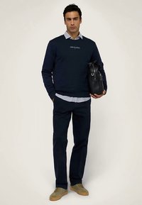 Mörkblå tröja med "TRUSSARDI"-logotyp, lager-på-lager över en ljusblå skjorta. Mörka byxor och beiga sneakers. Håller en svart väska.