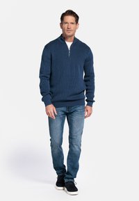Blauwe geribde ritstrui, casual pasvorm, gedragen over een witte shirt. Gecombineerd met lichtblauwe spijkerbroek en zwarte sneakers. Effen achtergrond.
