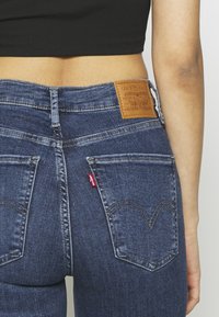 Jeans de mezclilla azul oscuro con cintura alta, que presentan un parche de cuero en la parte trasera de la cintura y costuras distintivas en los bolsillos traseros.
