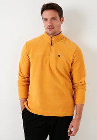 Žuti flis pulover s visokim ovratnikom i zatvaranjem na pola zatvarača. Ima mali crni logotip na prsima i duge rukave. Mekana tekstura.