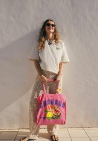 T-shirt oversize beige chiaro, pantaloni in pizzo bianchi e una borsa tote rosa con la scritta "GRANADA" e grafiche di frutta, su sfondo bianco.