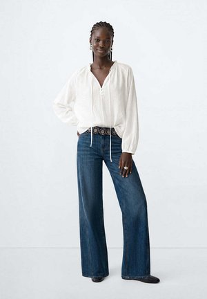 Femme aux cheveux tressés, debout, portant une blouse blanche à manches longues, un jean bleu à jambes larges, une ceinture noire avec des détails circulaires et des chaussures noires.
