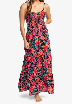 Femme portant une robe longue sans manches à motif floral avec des fleurs rouges, roses et orange et des feuilles vertes sur fond noir, debout pieds nus.