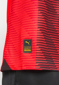 Maglia atletica rossa con motivo a zigzag nero, dotata di un'etichetta nera e dorata con la scritta "AUTENTICAMENTE LICENZIATA PER IL CALCIO" e un logo.