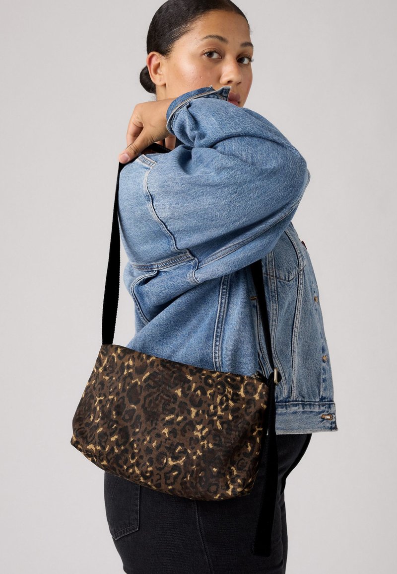 Vrouw in een lichtblauwe spijkerjacket en zwarte broek, met een bruine schoudertas in luipaardprint en een zwarte riem op haar rug.